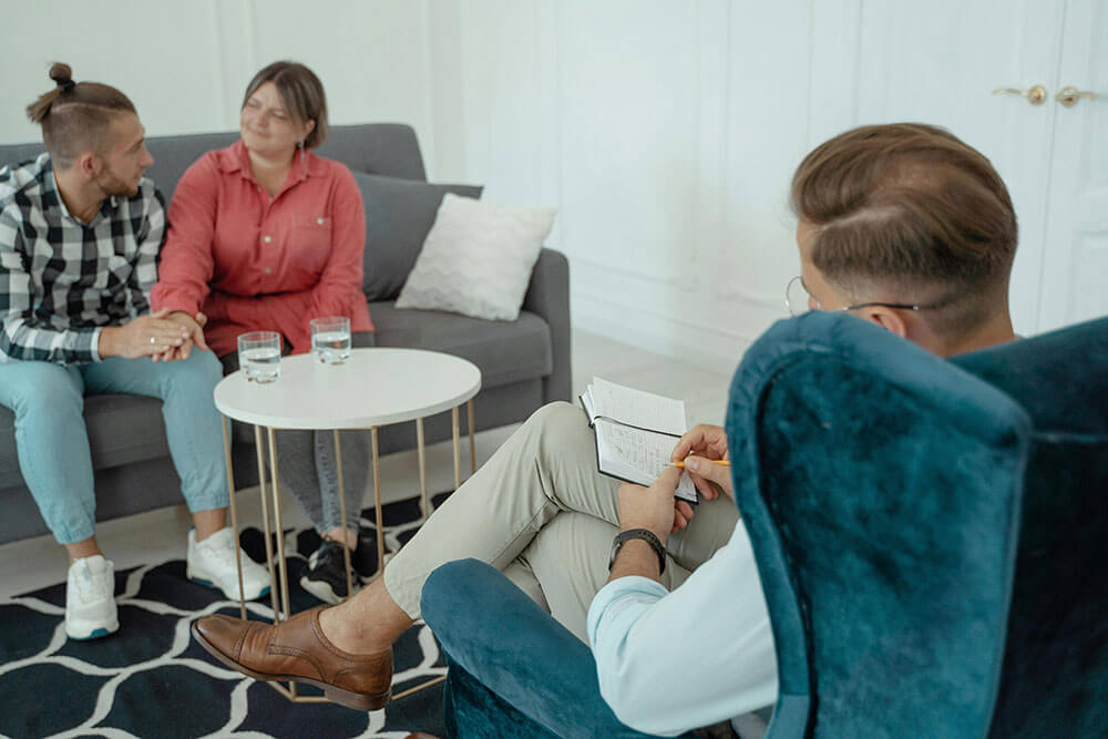 A couple sitting on a couch, holding hands, across from a man in a chair taking notes in a notebook