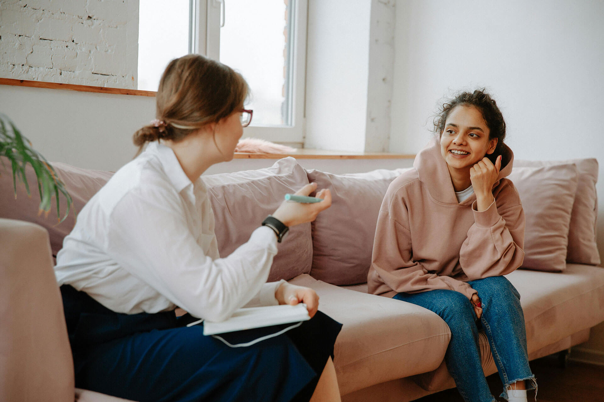 A professional woman with a notepad talking to another woman in a sweatshirt on a couch