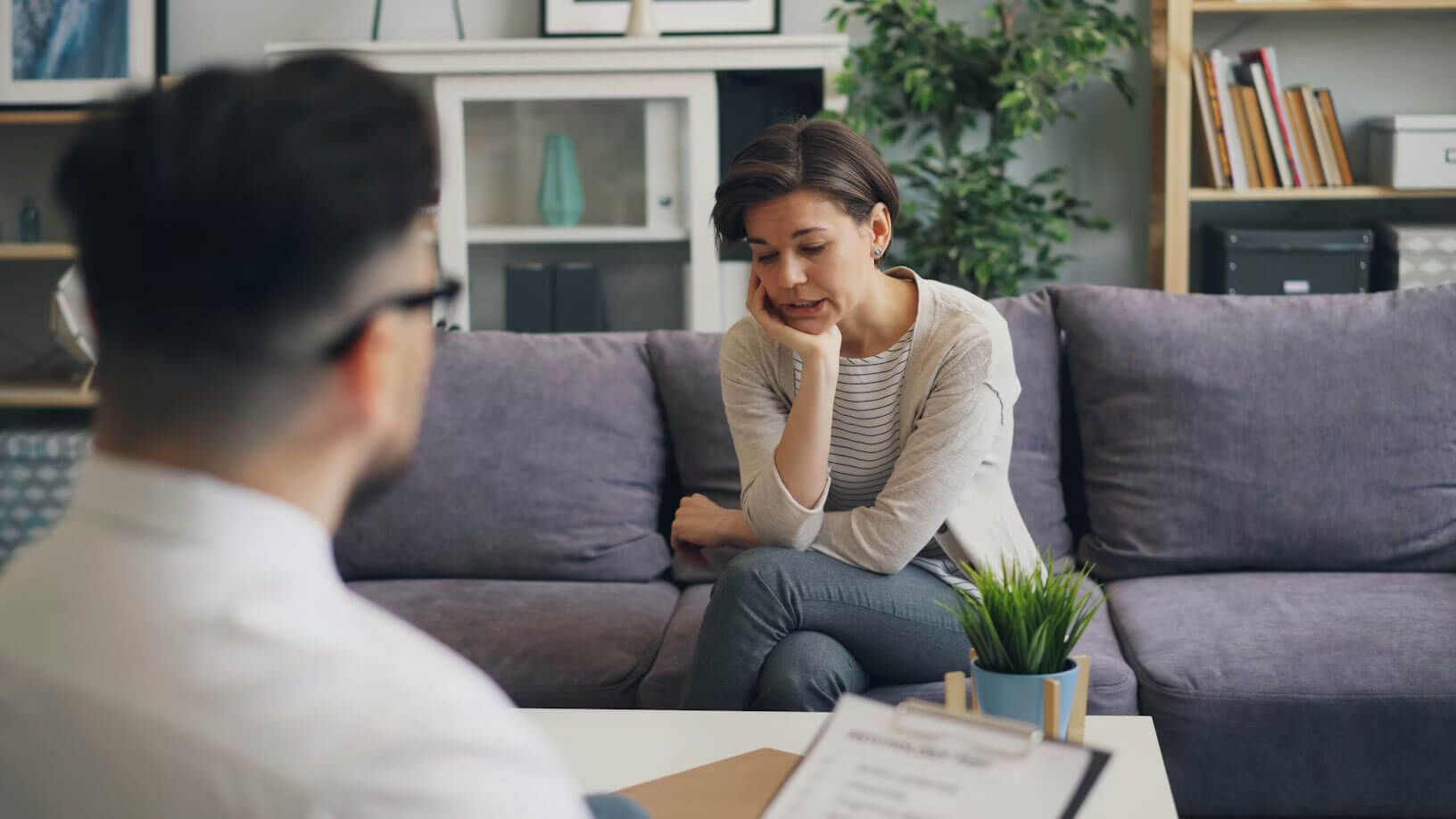 A person sitting on a couch across from a man holding a clipboard and wearing glasses