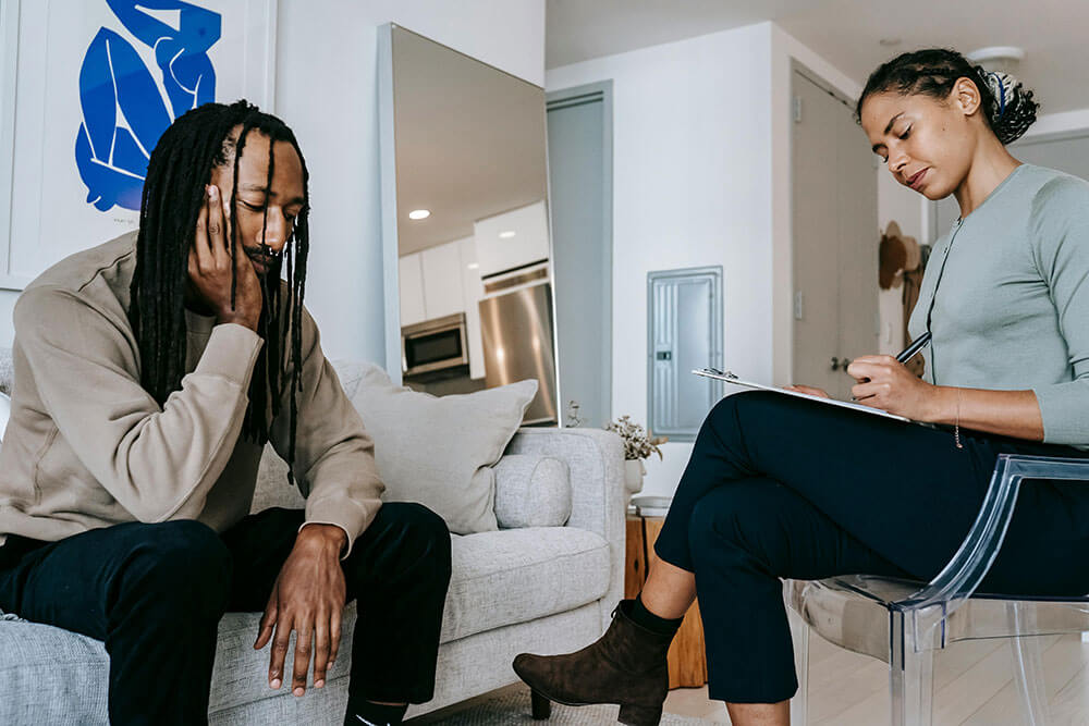 A somber looking man sitting on a couch across from a professional looking woman in a chair